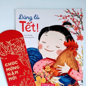 Peut inclure: Une enveloppe rouge et dorée avec le texte "CHÚC MUNG NAM MỚI" en lettres dorées. L'enveloppe est à côté d'un livre pour enfants avec une illustration colorée d'une fille tenant un coq. La couverture du livre porte le texte "Đúng là Tết!" en lettres noires.