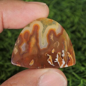May include: A brown, white and orange agate cabochon with a unique, swirling pattern. The cabochon is shaped like a teardrop and has a smooth, polished surface.