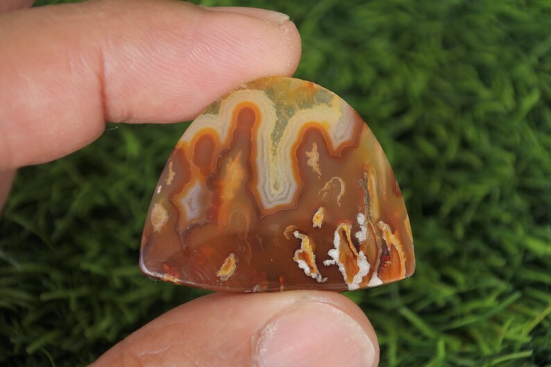 May include: A brown, white and orange agate cabochon with a unique, swirling pattern. The cabochon is shaped like a teardrop and has a smooth, polished surface.