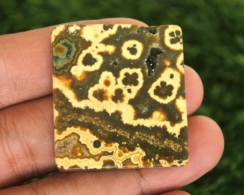 Puede incluir: Una piedra preciosa cuadrada con un patr&oacute;n &uacute;nico de manchas y remolinos amarillos, marrones y verde oscuro. La piedra tiene una superficie pulida y se sostiene en una mano, mostrando su belleza natural. La piedra preciosa probablemente se utiliza para joyer&iacute;a o fines decorativos.