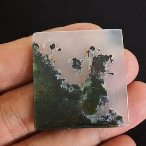 Puede incluir: Gema cuadrada con un patr&oacute;n de &aacute;gata musgosa. La piedra presenta una mezcla de colores verdes, blancos y marrones. La &aacute;gata musgosa tiene un dise&ntilde;o natural &uacute;nico. La piedra se sostiene en una mano sobre un fondo negro.