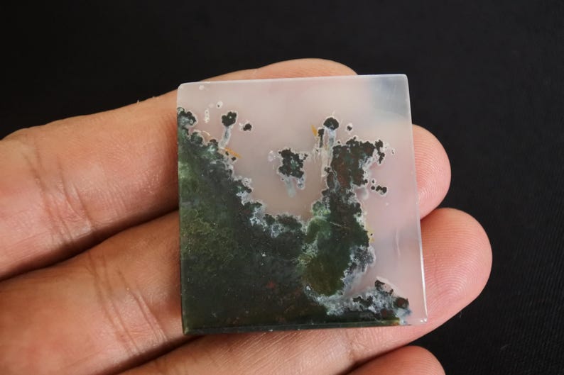 Puede incluir: Gema cuadrada con un patr&oacute;n de &aacute;gata musgosa. La piedra presenta una mezcla de colores verdes, blancos y marrones. La &aacute;gata musgosa tiene un dise&ntilde;o natural &uacute;nico. La piedra se sostiene en una mano sobre un fondo negro.