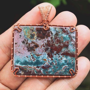 Puede incluir: Un colgante envuelto en alambre de cobre con una piedra de forma cuadrada que presenta un patrón moteado de marrón, rosa, azul y verde.