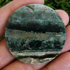 May include: A circular, polished stone displaying natural colours. The stone has a mix of dark green, teal, and brown tones, with a layered, banded design. The surface is glossy, emphasising the stone's unique veining and natural patterns.