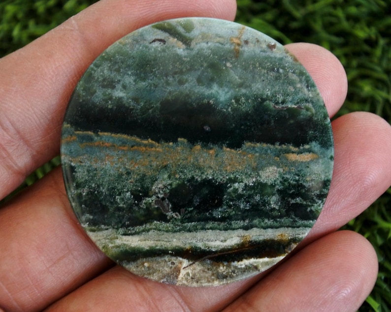 Op de afbeelding: Een ronde, gepolijste steen met een natuurlijk, aards uiterlijk. De steen heeft een mix van donkergroene, blauwgroene en bruine tinten, met een gelaagd, gestreept patroon. Het oppervlak heeft een glanzende afwerking, die de unieke adering van de steen benadrukt.