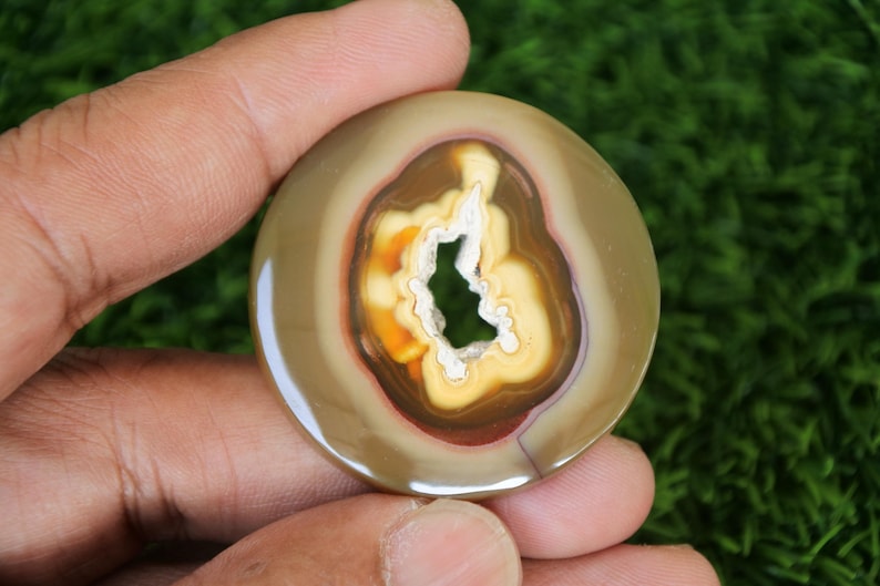 May include: A round, polished agate stone with a natural hole in the centre. The stone has a brown, tan, and white colour pattern.