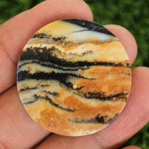 May include: A round, polished gemstone with a unique pattern of black, tan, and cream colors. The stone's surface features swirling lines and bands, creating a natural, abstract design. This gemstone is likely used for jewelry or decorative purposes.