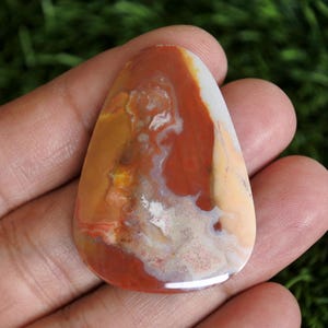 May include: A polished, teardrop-shaped cabochon gemstone featuring a marbled pattern of earthy tones including deep rust red, creamy beige, and translucent white. The stone has a smooth, glossy surface with visible natural inclusions and swirls.