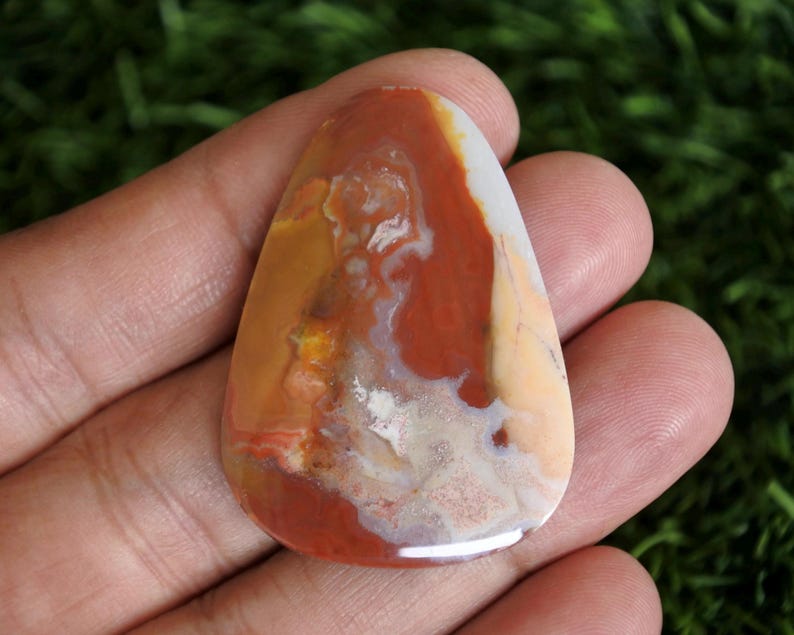 May include: A polished, teardrop-shaped cabochon gemstone featuring a marbled pattern of earthy tones including deep rust red, creamy beige, and translucent white. The stone has a smooth, glossy surface with visible natural inclusions and swirls.