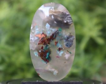 Puede incluir: Un cabujón ovalado transparente con inclusiones de cobre, turquesa y otros minerales. La piedra es translúcida y tiene un aspecto nublado.