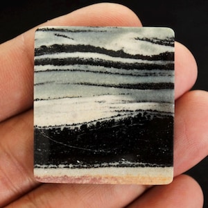 May include: A rectangular stone with a layered pattern of black, white, and grey bands. The stone has a polished surface and a light tan edge. The overall appearance resembles a landscape or abstract design.