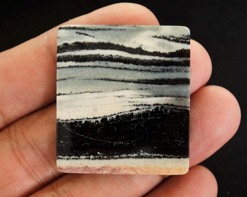 May include: A rectangular stone with a layered pattern of black, white, and grey bands. The stone has a polished surface and a light tan edge. The overall appearance resembles a landscape or abstract design.