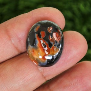 Op de afbeelding: Een ovaalvormige edelsteen met een gepolijst oppervlak. De steen heeft een mix van kleuren, waaronder zwart, oranje en wit, waardoor een uniek patroon ontstaat. De edelsteen wordt tussen de vingers gehouden, wat de grootte en vorm laat zien.