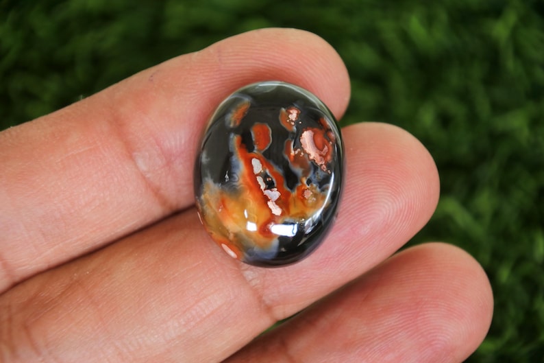 Może przedstawiać: Owalny kamień szlachetny o wypolerowanej powierzchni. Kamień ma mieszankę kolor&oacute;w, w tym czarny, pomarańczowy i biały, tworząc unikalny wz&oacute;r. Kamień szlachetny jest trzymany między palcami, pokazując jego rozmiar i kształt.
