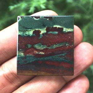 May include: A square gemstone with a layered pattern of deep red, green, and brown hues. The stone's surface has a polished appearance, revealing the natural variations in color and texture. The gemstone is held in a hand, with a blurred green background.