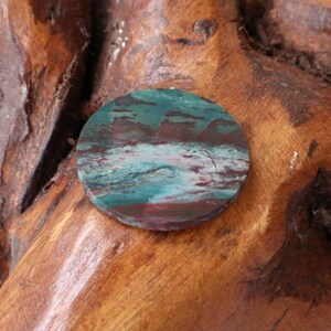 May include: A round, polished stone with a swirling pattern of green, brown, and white. The stone is set against a wooden background.