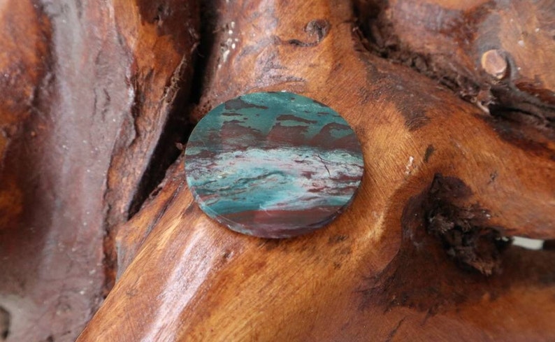 May include: A round, polished stone with a swirling pattern of green, brown, and white. The stone is set against a wooden background.