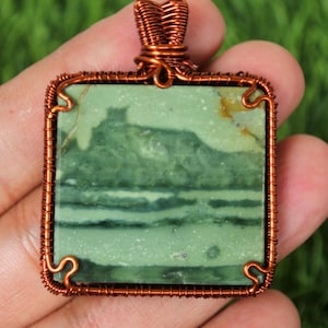 以下が含まれることがあります： 緑色の石を銅線で囲んだ正方形のペンダント。石は緑と茶色の色合いで風景のような模様があります。ワイヤーワークはフレームとネックレス用のベイルを形成しています。