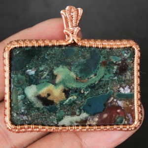 Może przedstawiać: Wisiorek owinięty miedzianym drutem z zielonym, brązowym i złotym agatem o wirującym wzorze.