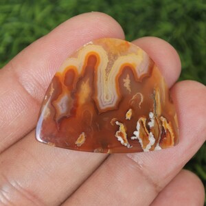 May include: A brown, orange, and white agate cabochon with a unique, swirling pattern. The cabochon is shaped like a teardrop and has a smooth, polished surface.