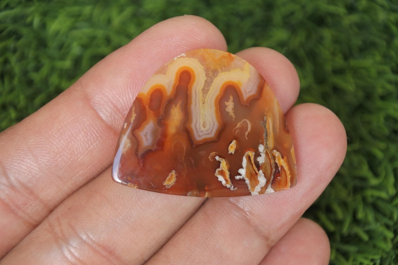 May include: A brown, orange and white agate cabochon with a unique, swirling pattern. The cabochon is shaped like a teardrop and has a smooth, polished surface.