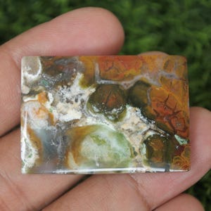 K&ouml;nnte beinhalten: Ein rechteckiger, polierter, mehrfarbiger Achat-Cabochon mit braunen, gr&uuml;nen, wei&szlig;en und orangefarbenen Mustern.