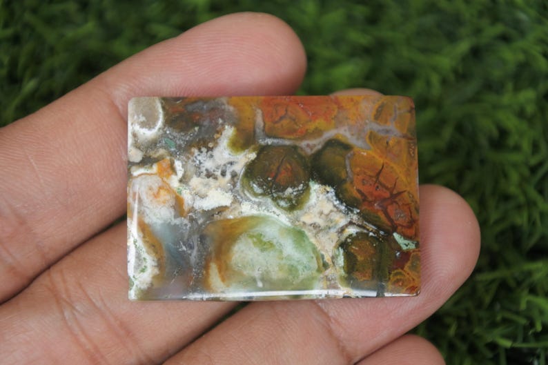 K&ouml;nnte beinhalten: Ein rechteckiger, polierter, mehrfarbiger Achat-Cabochon mit braunen, gr&uuml;nen, wei&szlig;en und orangefarbenen Mustern.