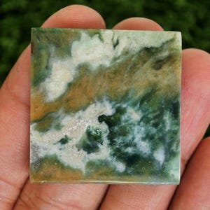 May include: Square gemstone with a natural pattern of green, white, and brown hues. The stone is held in a hand, showcasing its polished surface and unique colour variations. The gemstone's dimensions are approximately 3.8 cm by 3.8 cm.