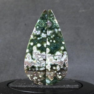 May include: Two polished gemstone cabochons, each with a teardrop shape. The stones feature a mix of green, white, and brown patterns, creating a unique and natural appearance. The cabochons are displayed on a black stand.