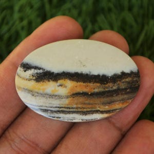 May include: A white, brown and black banded oval cabochon gemstone. The stone has a layered pattern with a white top layer and brown and black bands underneath.