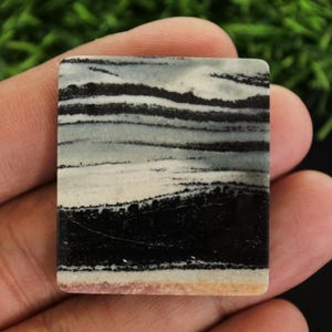 May include: A rectangular stone with a layered pattern of black, white, and grey bands. The stone is held in a hand, showcasing its polished surface and natural colour variations. The bottom edge has a light tan hue.