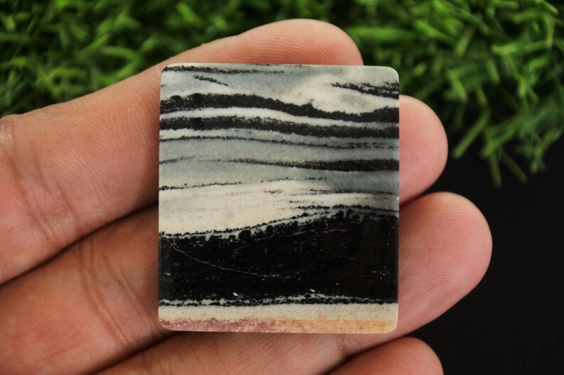 May include: A rectangular stone with a layered pattern of black, white, and grey bands. The stone is held in a hand, showcasing its polished surface and natural colour variations. The bottom edge has a light tan hue.