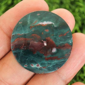 Peut inclure: Une pierre pr&eacute;cieuse ronde et polie, m&eacute;langeant des teintes de vert-bleu profond et de bordeaux. La pierre pr&eacute;sente un motif tourbillonnant avec des veines blanches et rouges. Elle est tenue dans une main, sur un fond vert flou.