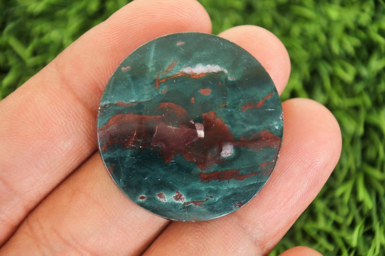 Peut inclure: Une pierre pr&eacute;cieuse ronde et polie, m&eacute;langeant des teintes de vert-bleu profond et de bordeaux. La pierre pr&eacute;sente un motif tourbillonnant avec des veines blanches et rouges. Elle est tenue dans une main, sur un fond vert flou.