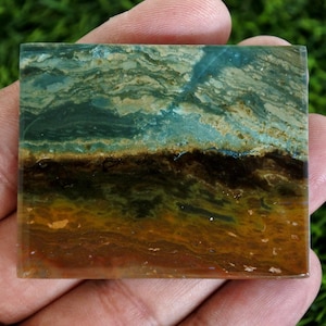 May include: A square, polished stone with a layered appearance. The top section has swirling patterns of teal and tan, leading to a dark brown band, and then to a golden-brown base. The stone is held in a hand against a green background.