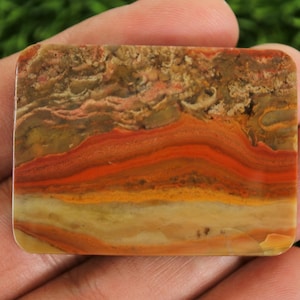 May include: A rectangular gemstone with a layered pattern of orange, red, yellow, and brown hues. The stone is held in a hand, showcasing its polished surface and natural color variations. The gemstone's design resembles a landscape.