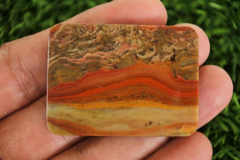 Puede incluir: Una gema rectangular con un patr&oacute;n en capas de tonos naranja, rojo, amarillo y marr&oacute;n. La piedra se sostiene en una mano, mostrando su superficie pulida y variaciones de color naturales. El dise&ntilde;o de la gema se asemeja a un paisaje.