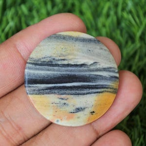 Puede incluir: Una piedra redonda y pulida con un patrón de bandas de negro, blanco y amarillo. La piedra tiene una superficie lisa y pulida.