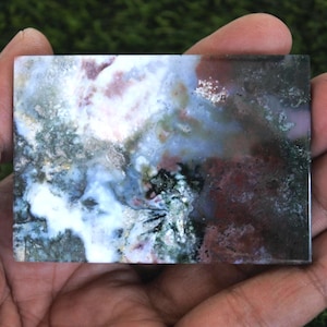 K&ouml;nnte beinhalten: Ein rechteckiger, polierter Stein mit einem nat&uuml;rlichen, abstrakten Muster. Der Stein weist eine Mischung aus Farben auf, darunter Wei&szlig;, Blau, Gr&uuml;n und Kastanienbraun. Der Stein wird in einer Hand gehalten, um seine Gr&ouml;&szlig;e und Textur zu zeigen. Die Gesamt&auml;sthetik ist erdig und organisch.