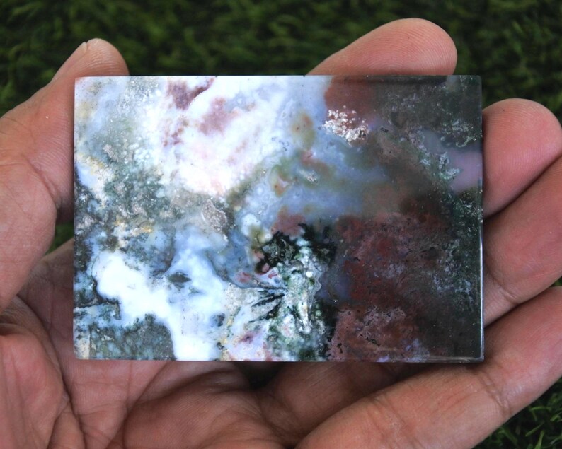 K&ouml;nnte beinhalten: Ein rechteckiger, polierter Stein mit einem nat&uuml;rlichen, abstrakten Muster. Der Stein weist eine Mischung aus Farben auf, darunter Wei&szlig;, Blau, Gr&uuml;n und Kastanienbraun. Der Stein wird in einer Hand gehalten, um seine Gr&ouml;&szlig;e und Textur zu zeigen. Die Gesamt&auml;sthetik ist erdig und organisch.