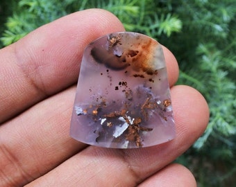 May include: A polished, translucent gemstone with a unique, organic pattern. The stone features a blend of lavender, brown, and white hues, with intricate, branching inclusions. The top is rounded, and the bottom is a flat, curved edge. The gemstone is held in a hand.