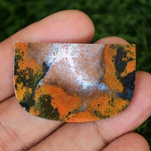 May include: A semi-circular gemstone with a polished finish, displaying a distinctive pattern. The stone's colours include orange, black, white, and green, forming a natural, abstract design. The gemstone is held in a hand, highlighting its size and detail.