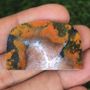 May include: A semi-circular gemstone with a polished finish, displaying a distinctive pattern. The stone's colours include orange, black, white, and green, forming a natural, abstract design. The gemstone is held in a hand, highlighting its size and texture.