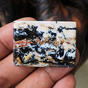 May include: A rectangular, white, black and brown agate cabochon with a unique, abstract pattern. The stone has a polished surface and is approximately 5 cm long.