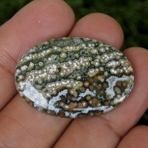 May include: Oval gemstone with a distinctive pattern. The stone displays a blend of green, brown, and white hues, giving a speckled look. The polished surface reflects light, accentuating the natural variations. Suitable for jewellery making, this stone is a unique addition.
