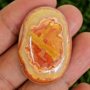 Peut inclure: Pierre précieuse ovale polie avec un motif vibrant de couleurs jaune, orange et rouge. La surface de la pierre présente un motif abstrait en couches, rappelant un paysage. La pierre est tenue dans une main, mettant en valeur sa taille et sa forme.