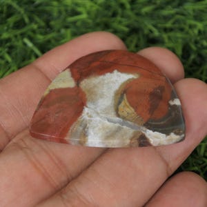Op de afbeelding: Een rode, witte en bruine agaat cabochon met een uniek, draaiend patroon. De cabochon is gevormd als een gitaarplectrum.