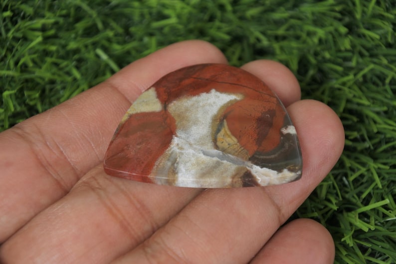 Op de afbeelding: Een rode, witte en bruine agaat cabochon met een uniek, draaiend patroon. De cabochon is gevormd als een gitaarplectrum.