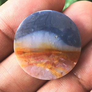 Peut inclure: Une pierre ronde et polie avec un motif de paysage. La partie supérieure est bleu foncé, passant au blanc, à l'orange et au brun rougeâtre en bas. La pierre a une finition lisse et brillante.