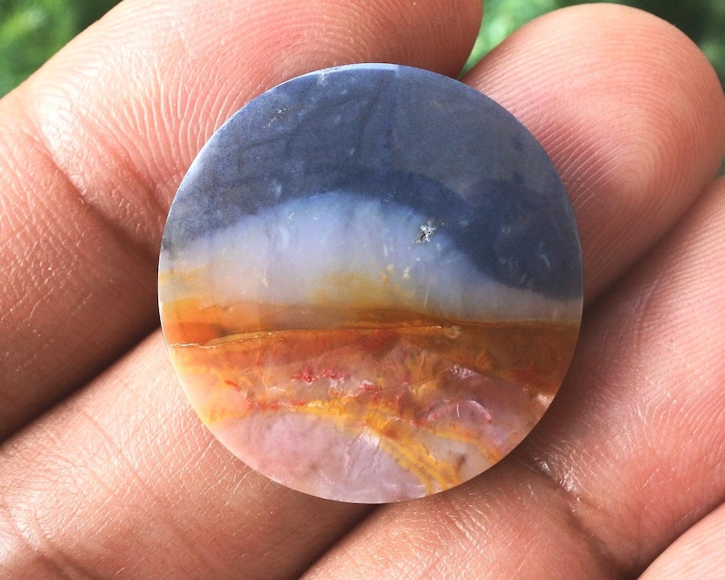 May include: A circular, polished stone displaying a landscape pattern. The top section is a deep blue, fading into white, orange, and reddish-brown tones towards the base. The stone has a smooth, glossy finish.